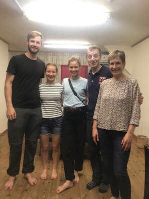 Eine Gruppe von Personen steht in einem Raum mit Holzboden und einer schlichten Wand im Hintergrund.