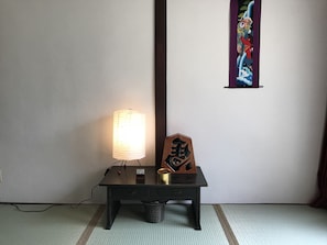 Um quarto com piso de tatame, uma luminária tradicional japonesa, um pequeno armário de madeira com caligrafia e um quadro colorido na parede.