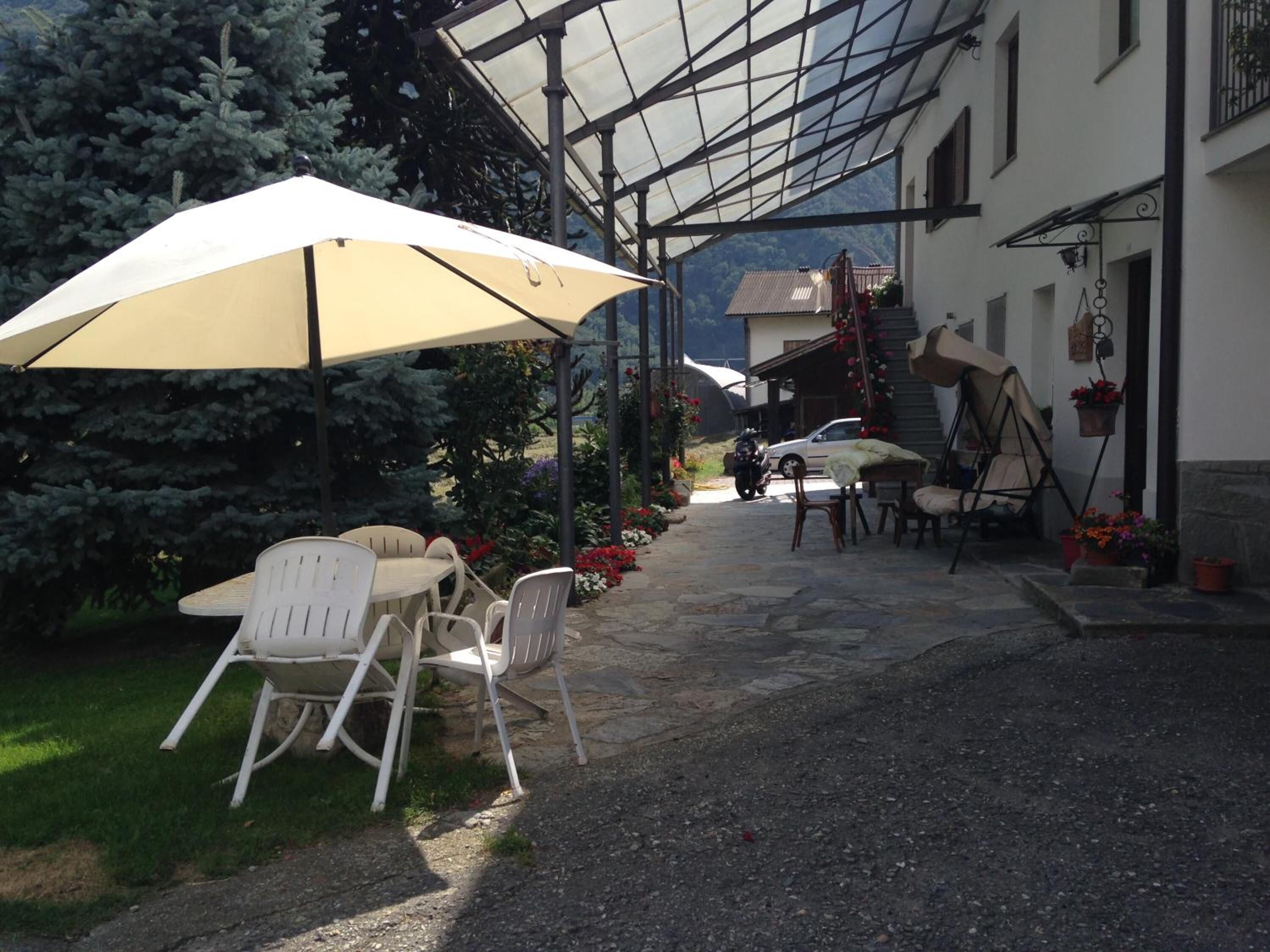 A covered outdoor seating area with chairs and a table, a motorcycle parked in the background, and a building with a staircase.