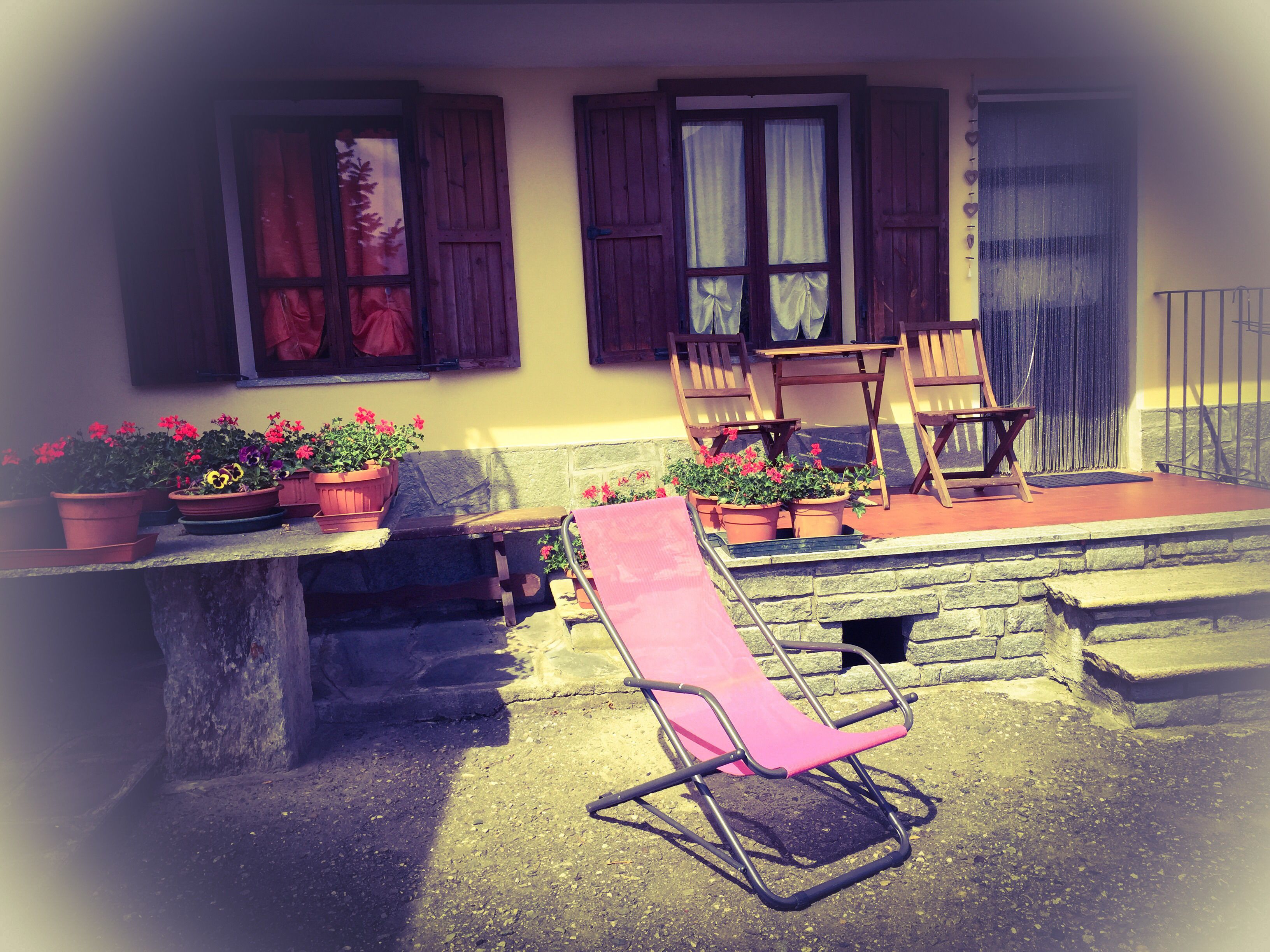 A stone bench with a pink chair in front, wooden chairs, and potted plants on a balcony.