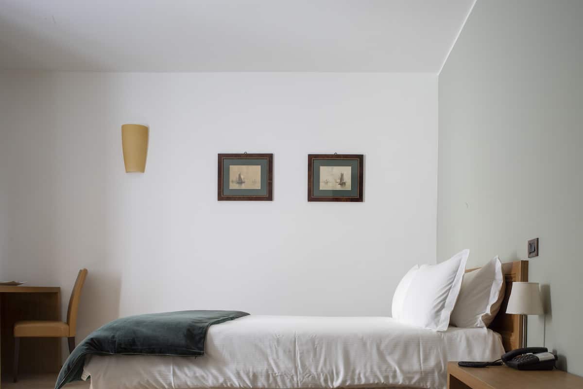 Ein Hotelzimmer mit einem Bett, einem Schreibtisch, einem Stuhl, zwei gerahmten Bildern an der Wand, einer Lampe und einem Telefon auf dem Nachttisch.