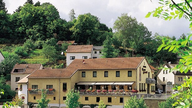 A riverside hotel with a terracotta roof, a balcony with flowers, and a view of the river.
