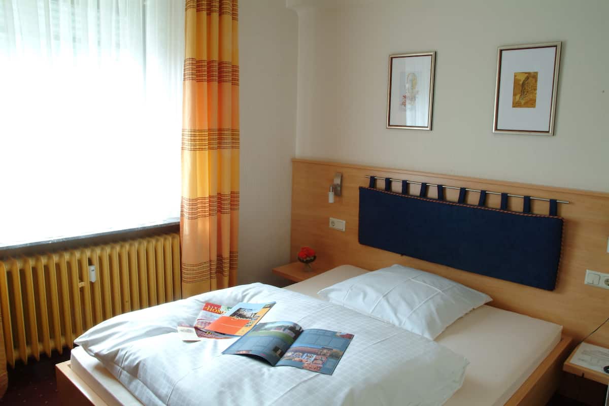 Una habitación de hotel con una cama, una mesita de noche, un ventanal con cortinas y dos cuadros en la pared.