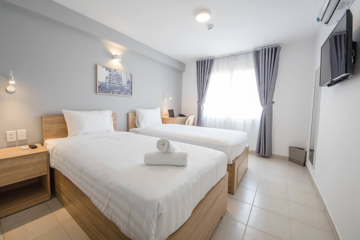 A hotel room with two beds, a flat-screen TV, a painting on the wall, and a window with curtains.