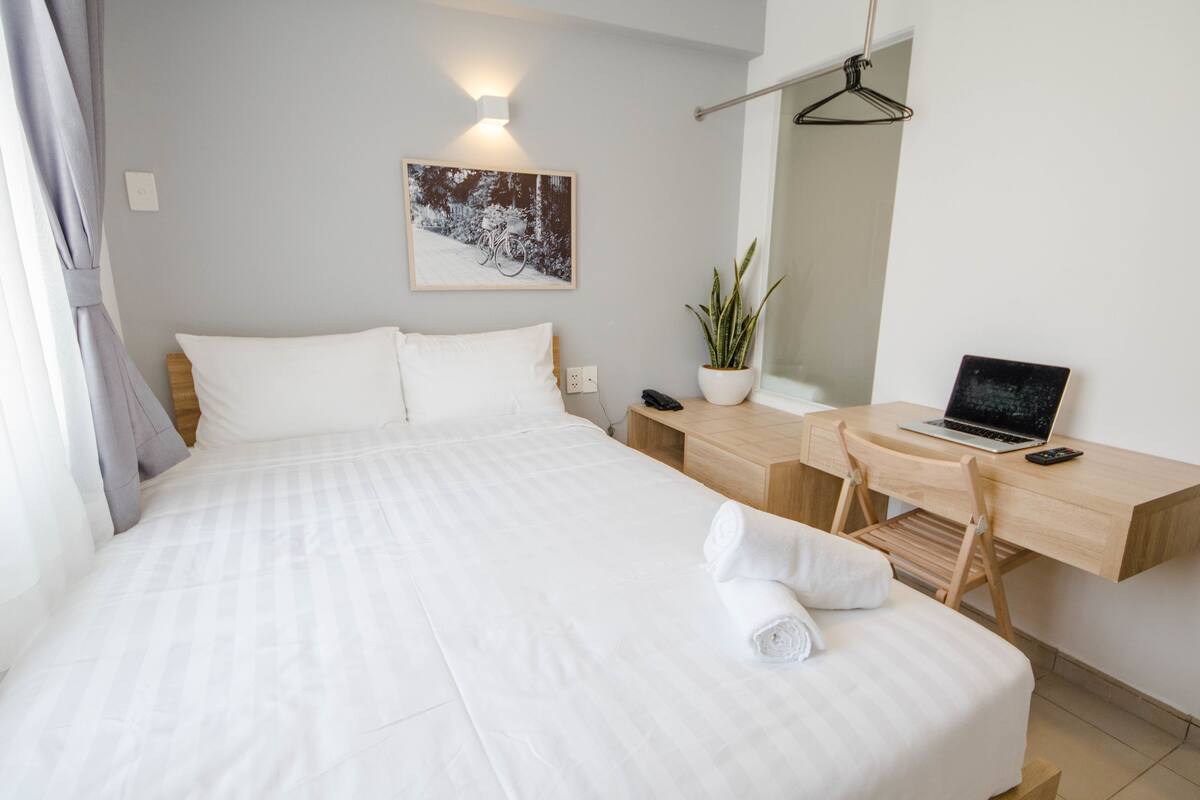 A neatly made bed with white linens, a wooden desk with a laptop, a potted plant, and a wall-mounted mirror.