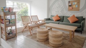 A modern living room with a green sofa, wooden chairs, a wooden table, and wicker ottomans. There is a shelf with snacks and a wall map. - COMMON INN Thao Dien (Ho Chi Minh City)