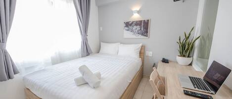 Un lit bien fait, avec du linge de lit blanc, un ordinateur portable posé sur un bureau en bois, une plante en pot et un luminaire fixé au mur.