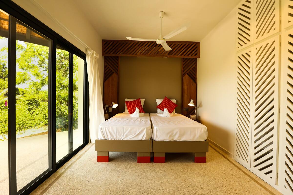 A double bed with a ceiling fan, large windows, and a view of greenery outside.
