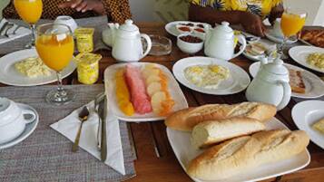 Una mesa de desayuno con platos de comida, entre ellos huevos, tostadas y fruta, junto con una tetera y tazas.