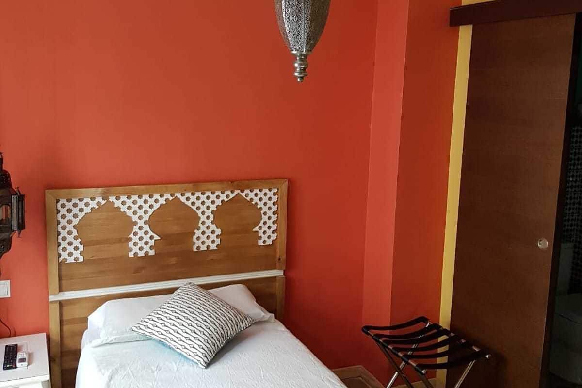 A room with a bed, a hanging light fixture, a wooden headboard, a chair, and a small table with a remote control.