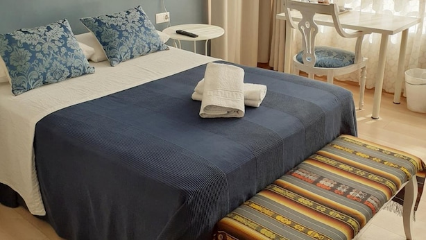 Un dormitorio con cama, mesita de noche, lámpara, silla y un banco.