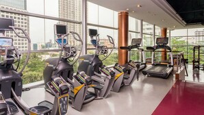 A gym with treadmills, ellipticals, and stationary bikes, large windows, and a red floor.