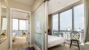 A modern hotel room with a large bed, a chair, and a view of the city through large windows.