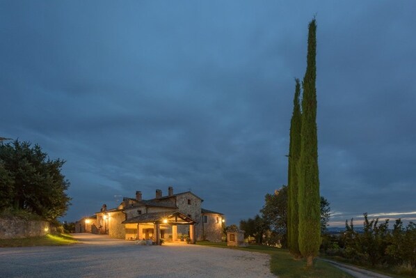 A stone house with a gravel driveway, a tall cypress tree, and a view of the countryside.