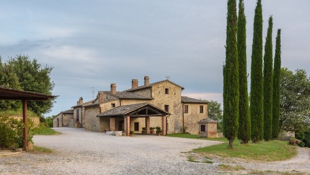 A stone building with a gravel driveway, surrounded by tall cypress trees. - Agriturismo Cerreto (Pienza)
