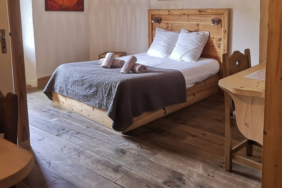 Un lit en bois avec des draps blancs et une couverture foncée, une table en bois et un luminaire suspendu au plafond.