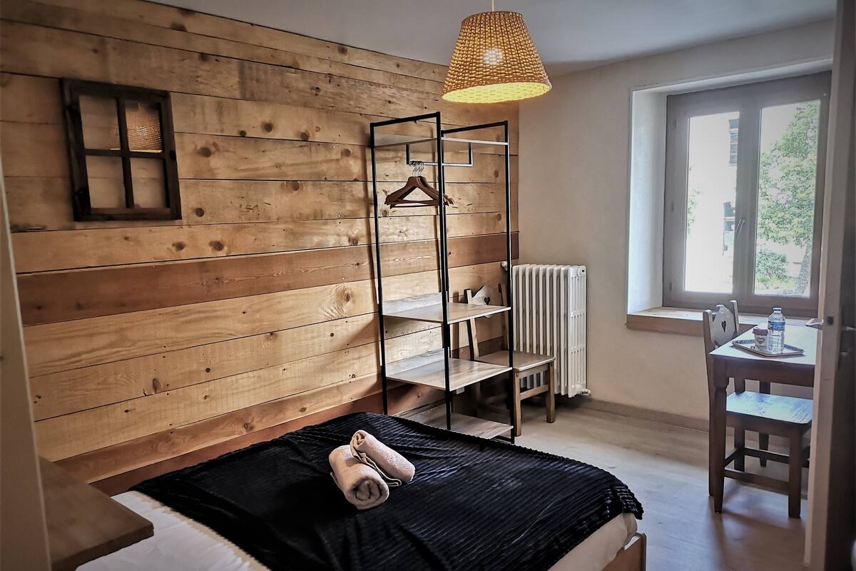 Une chambre avec une tête de lit en bois, un luminaire suspendu, un radiateur, un bureau avec une chaise et une fenêtre donnant sur un paysage verdoyant.