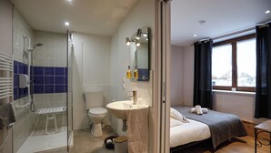 A modern bathroom with a glass shower enclosure, a toilet, and a sink with a mirror above it. There is a bed with a dark-colored blanket and pillows, and a window with curtains. - Auberge Les 2 Moucherolles (Correncon-en-Vercors)
