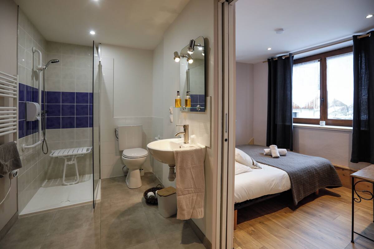 Une salle de bain moderne avec une douche à parois de verre, des toilettes et un lavabo avec un miroir au-dessus. On y trouve un lit avec une couverture et des oreillers de couleur foncée, ainsi qu’une fenêtre avec des rideaux.
