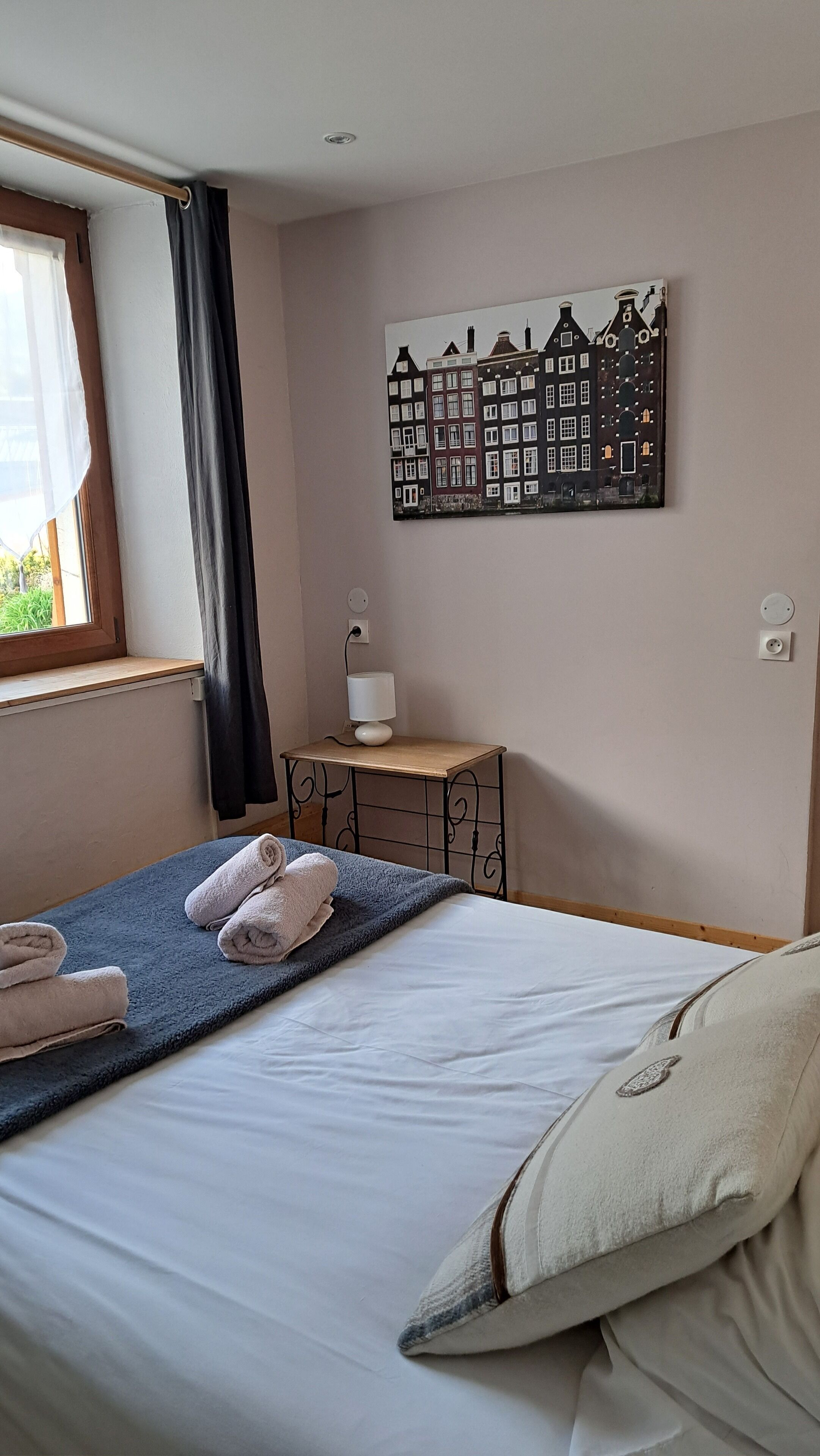 A bedroom with a bed, towels, a lamp, and a wall art of buildings.