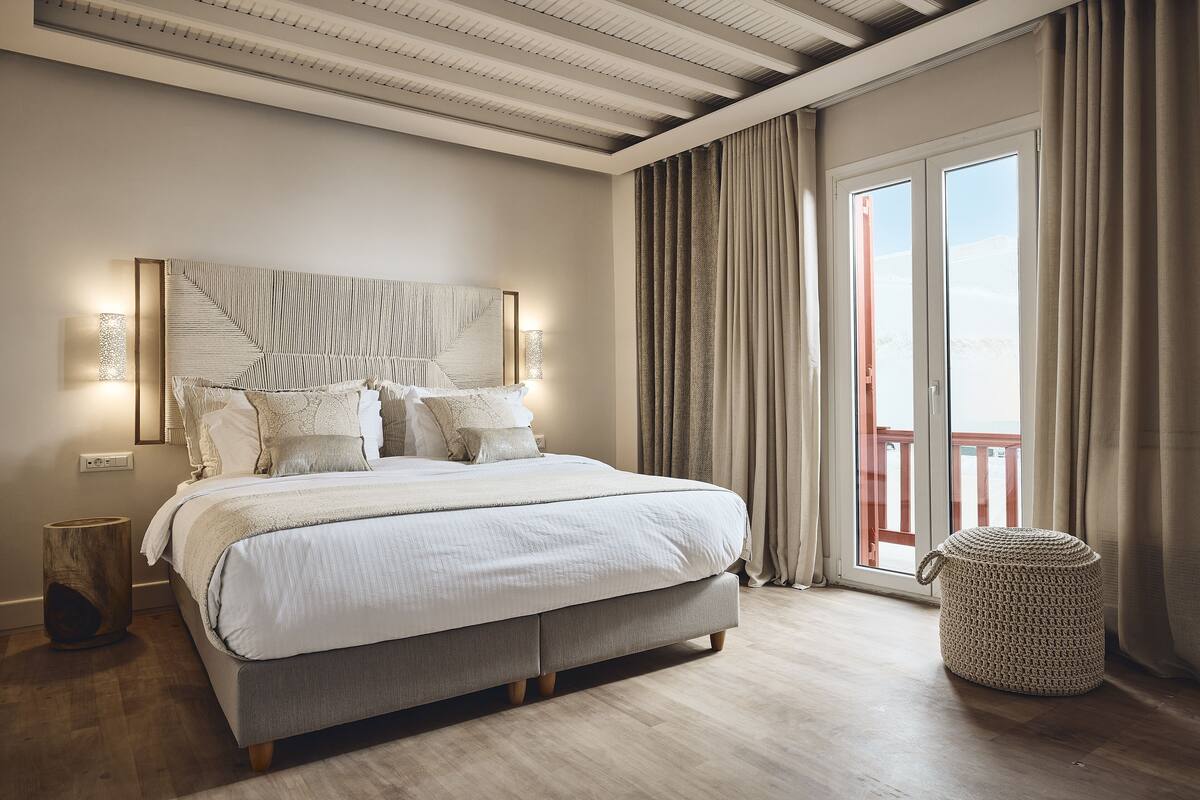 A bedroom with a large bed, a bedside lamp, a wooden floor, and a view of the outdoors through a sliding door.