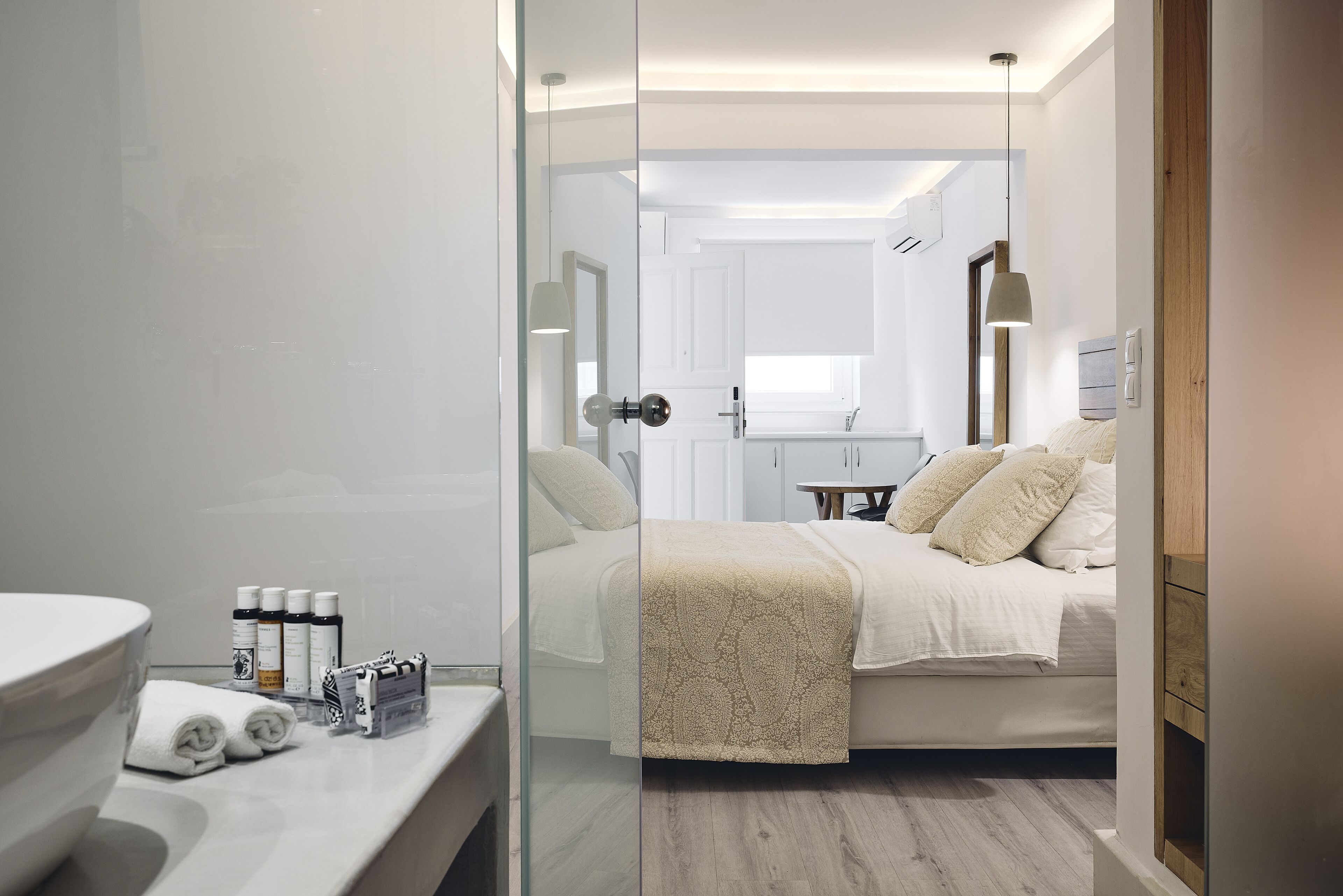 Ett modernt hotellrum med en glasvägg, en säng med beige sängkläder, ett sängbord med toalettartiklar och utsikt över det intilliggande badrummet.
