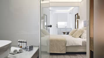Ett modernt hotellrum med en glasvägg, en säng med beige sängkläder, ett sängbord med toalettartiklar och utsikt över det intilliggande badrummet.