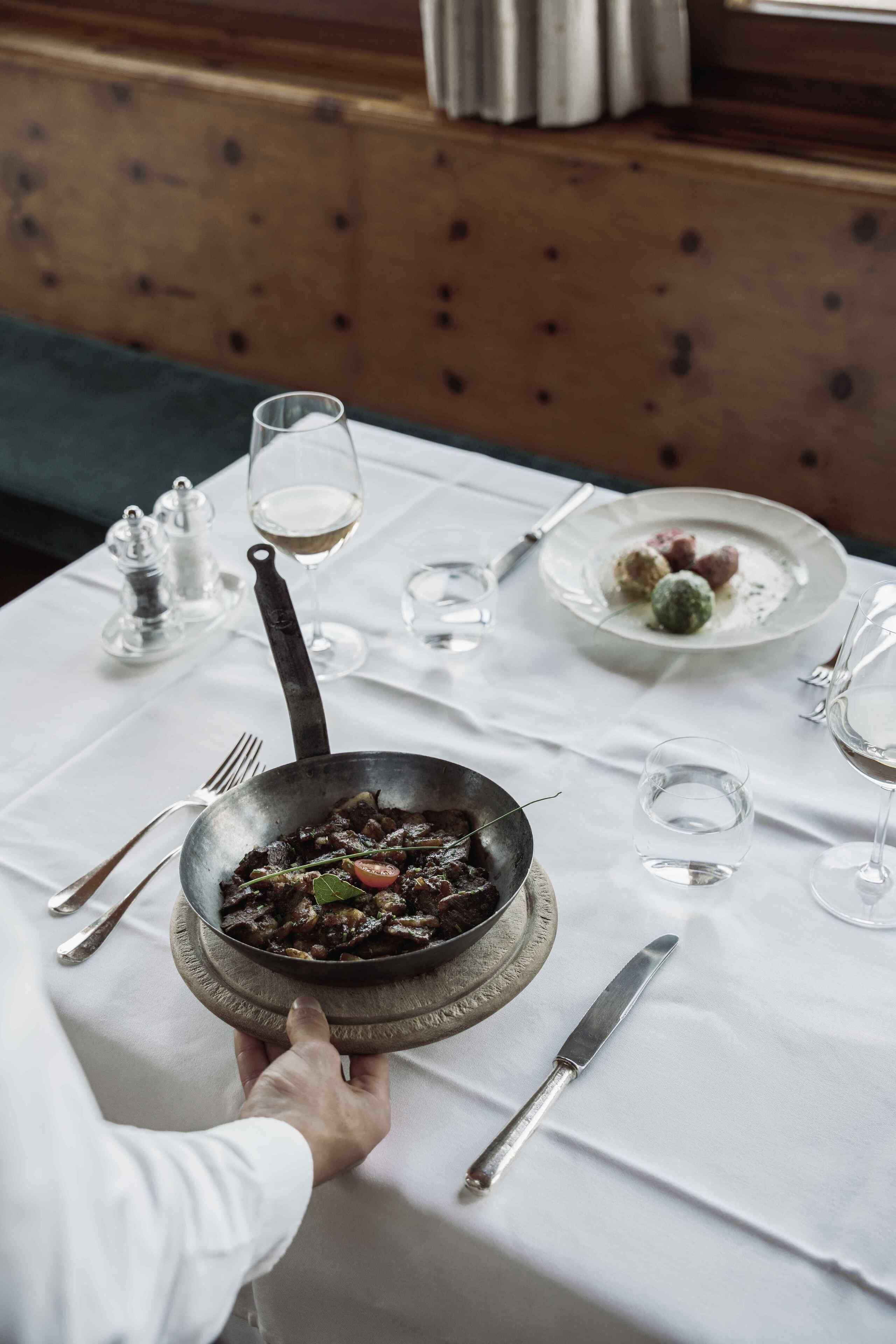 Een eettafel gedekt met een braadpan eten, wijnglazen en bestek.