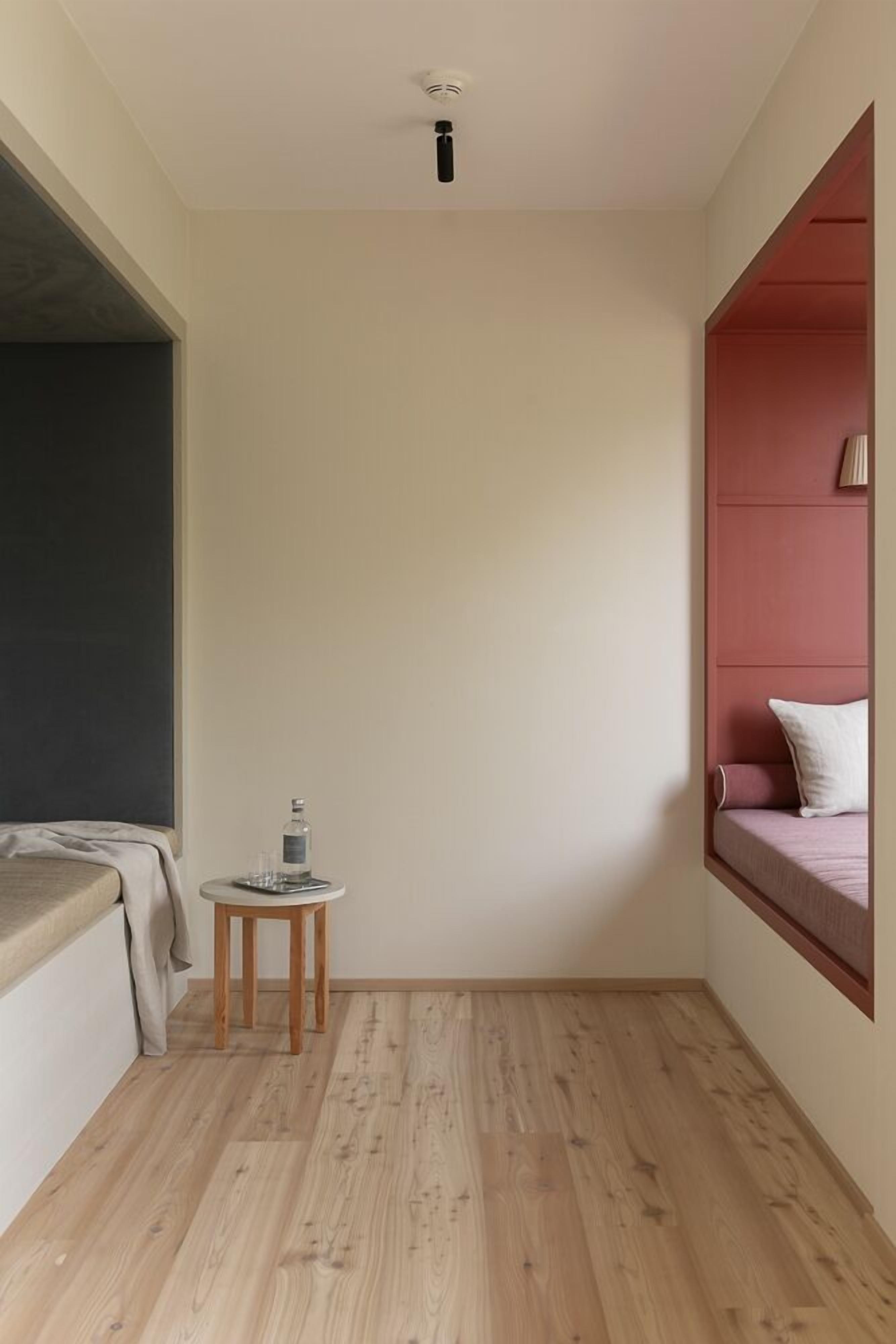 Una habitación pequeña y moderna con cama, una mesita con una botella y vasos, y una ventana con pared roja.