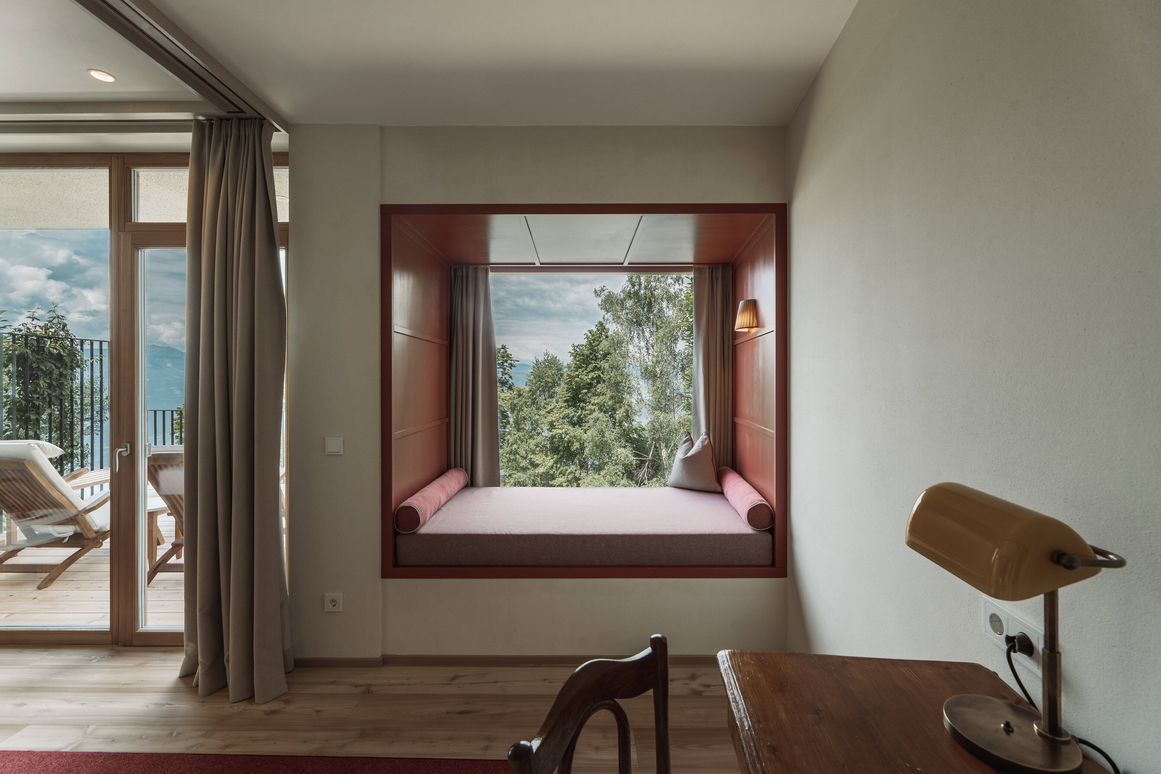 Ein Zimmer mit eingebautem Fensterbrett, einem Schreibtisch mit Lampe und Blick auf einen Balkon mit Gartenmöbeln.