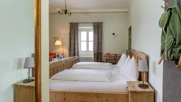 A wooden bed with white linens, two bedside tables with lamps, a window with curtains, a radiator, and a coat hanging on the wall.