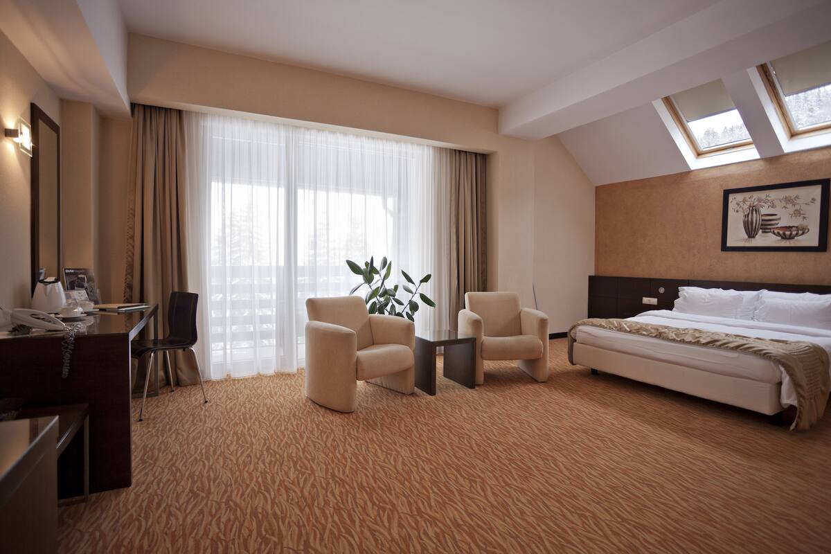 Une chambre d’hôtel comprenant un lit, deux fauteuils, un bureau avec un téléphone et une grande fenêtre avec des rideaux.