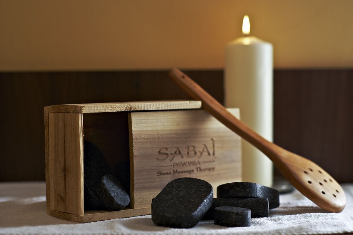 Una caja de madera con la etiqueta "SABAI", piedras negras, una espátula de madera y una vela encendida sobre una mesa.