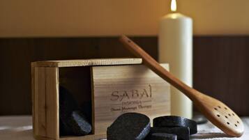 Una caja de madera con la etiqueta "SABAI", piedras negras, una espátula de madera y una vela encendida sobre una mesa.