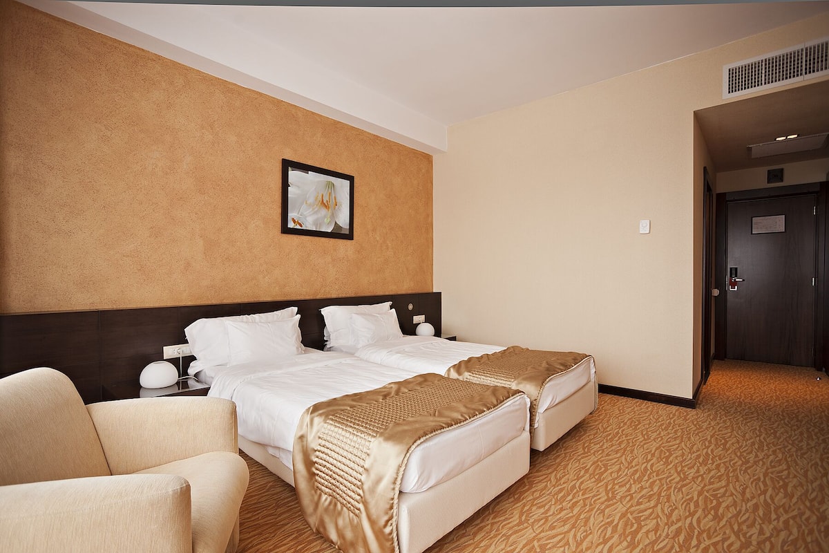 Une chambre d’hôtel avec un lit double, deux tables de chevet, une chaise, un grand tableau au mur et une porte donnant sur une autre pièce.