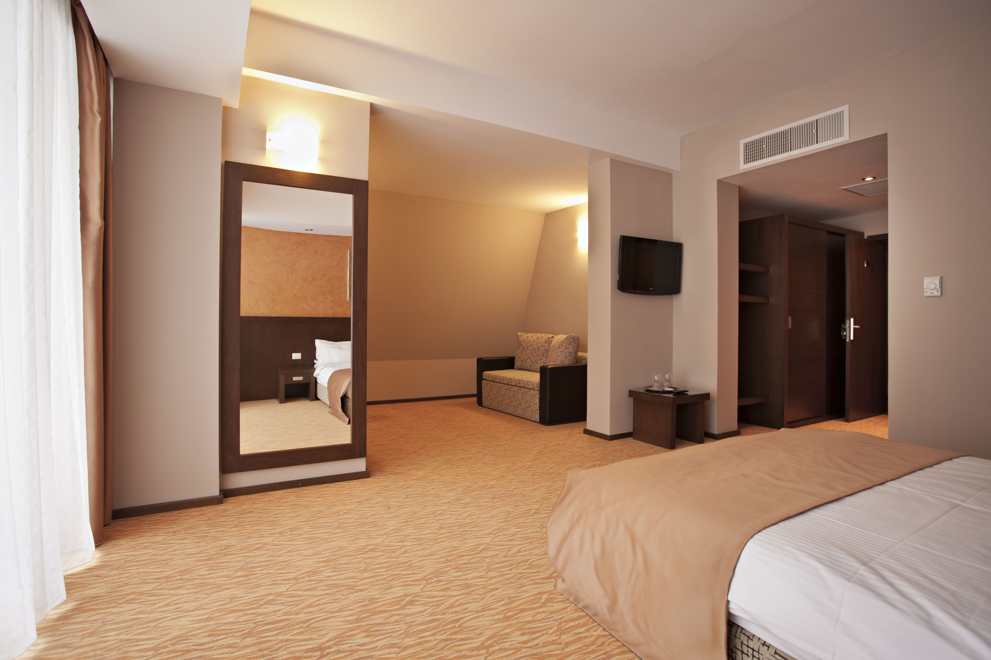 A hotel room with a large bed, a sitting area with a chair and a small table, a mirror, a flat-screen TV mounted on the wall, and a wardrobe.
