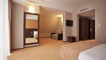 A hotel room with a large bed, a sitting area with a chair and a small table, a mirror, a flat-screen TV mounted on the wall, and a wardrobe.
