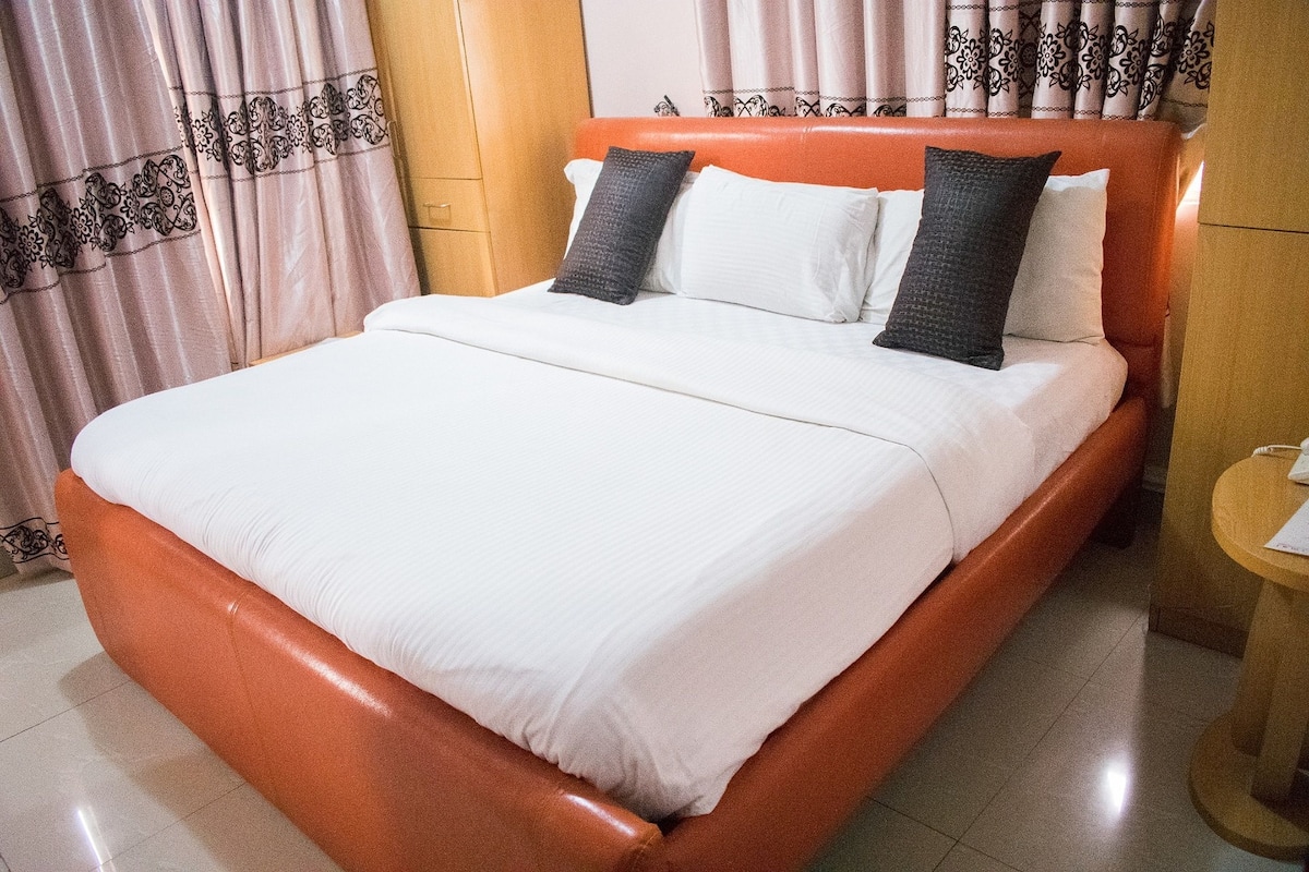 A neatly made bed with white bedding and two dark pillows, a wooden headboard, and a small wooden side table with a phone.