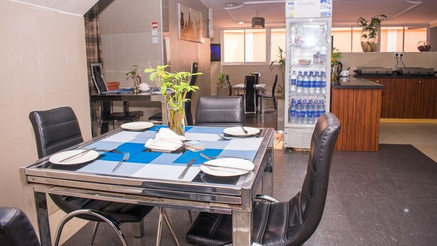 A dining area with a glass table set for four, surrounded by black leather chairs. A refrigerator with bottled water is visible in the background. - Primal Hotel (Lagos)