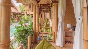 A bamboo-framed room with a thatched roof, a bed with a green pillow, and a view of a pool and greenery outside. - Moon Bamboo (Pererenan)