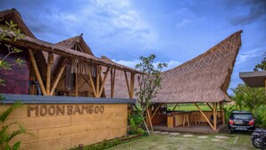 A traditional bamboo structure with a thatched roof, a sign reading "MOONBAMBOO", and a paved entrance area with a car and motorcycle parked nearby. - Moon Bamboo (Pererenan)