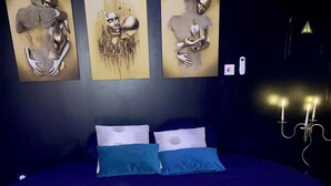 A large bed with a dark blue bedspread and two pillows, a wall with three abstract art pieces, and a wall-mounted light fixture. - Le Gambetta (Vias)