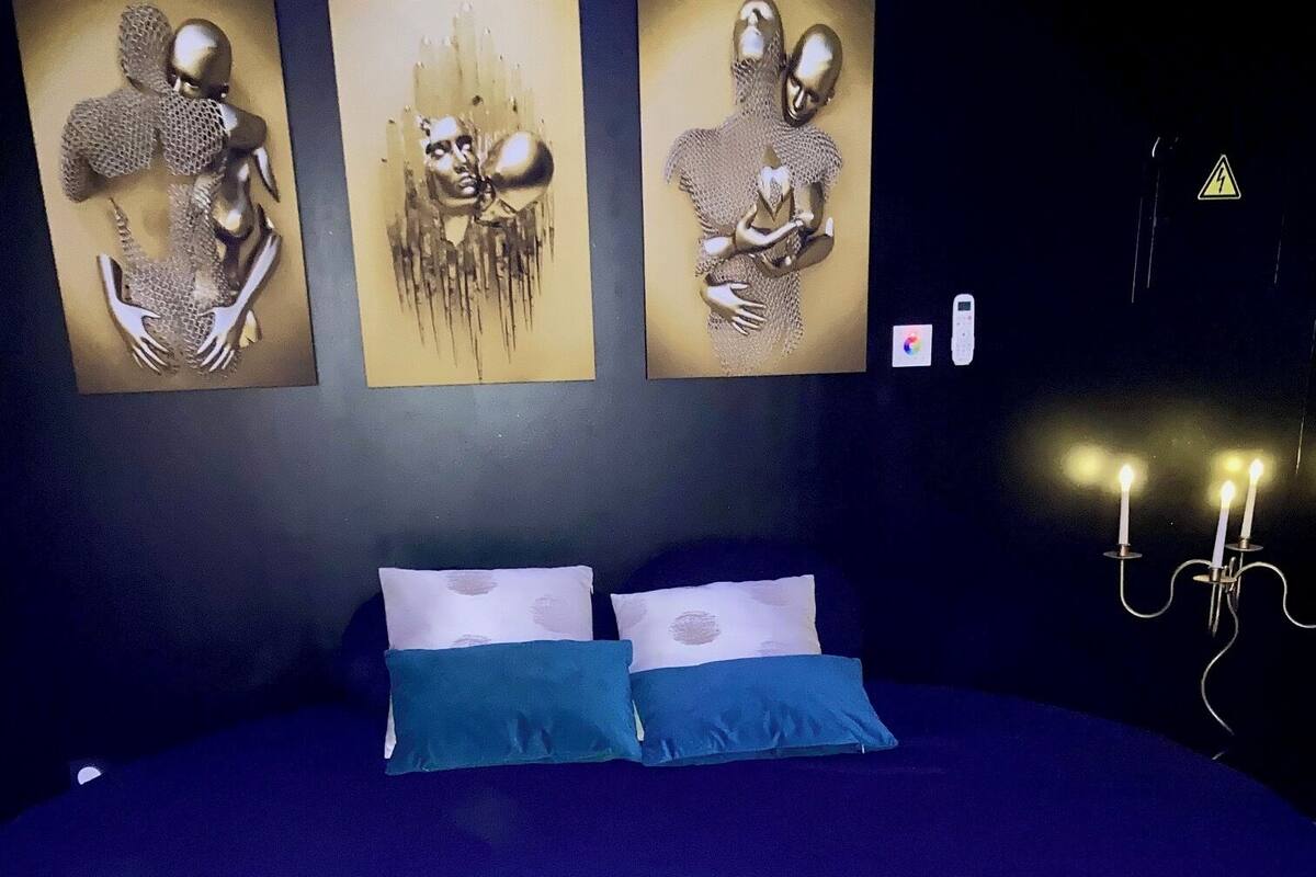 A large bed with a dark blue bedspread and two pillows, a wall with three abstract art pieces, and a wall-mounted light fixture.