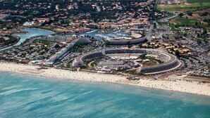 An aerial view of a coastal area with a large circular building, likely a stadium or arena, surrounded by a marina with boats. - Le Gambetta (Vias)