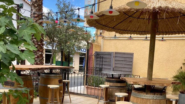 An outdoor patio with wooden barrels as tables and stools, surrounded by potted plants and a palm tree. - Le Gambetta (Vias)