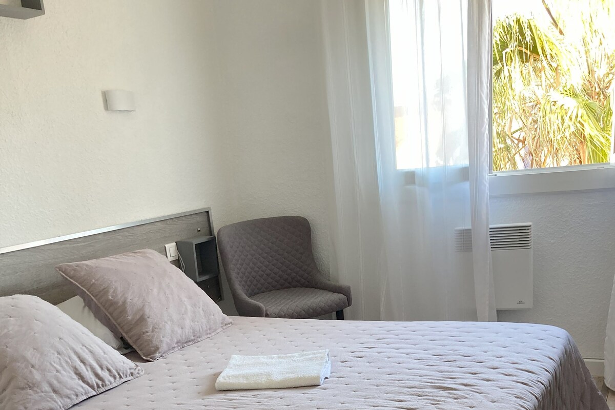 Un lit bien fait, recouvert d’une courtepointe, une chaise et une table de chevet dans une pièce dotée d’un plancher en bois et d’une fenêtre avec des rideaux.