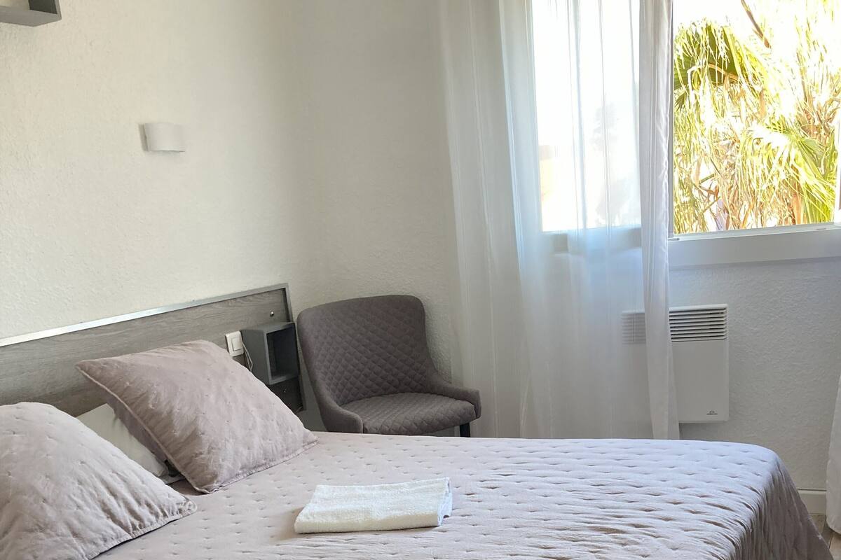 A neatly made bed with a quilted cover, a chair, and a bedside table in a room with a wooden floor and a window with curtains.