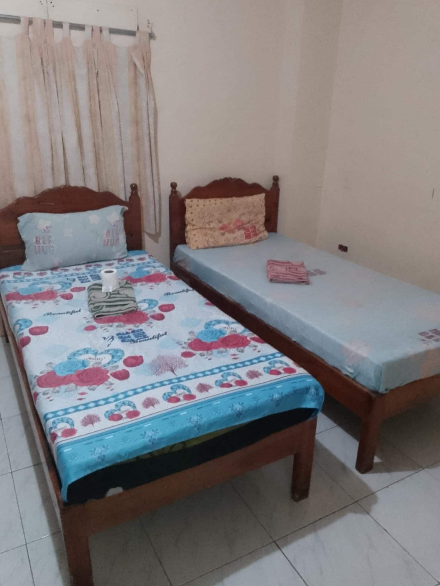 A room with two single beds, one with a floral bedspread and the other with a plain one. There is a wooden headboard on one bed and a curtain on the window.