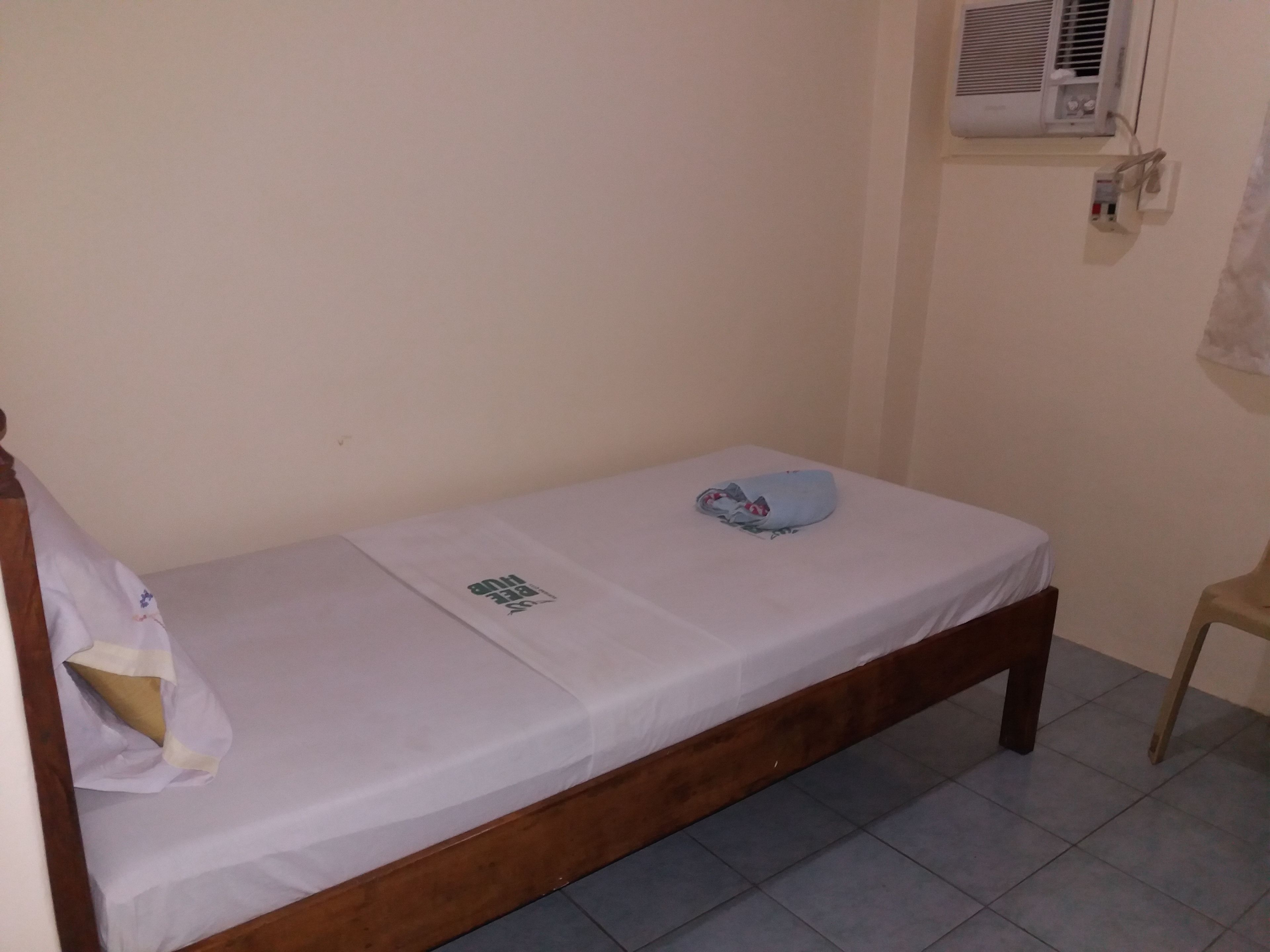 Una cama individual con estructura de madera, colchón blanco y manta azul. Arriba de la cama, hay un equipo de aire acondicionado instalado en la pared.