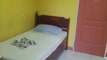 Una habitación pequeña con una cama individual, cabecera de madera, colchón blanco y almohada azul. Se observa una puerta roja con un aviso, y las paredes son amarillas.